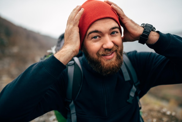 屋外は山でハイキングの赤い帽子をかぶった幸せなハイカーの若い男の肖像画をクローズアップ旅行者のひげを生やした男性の笑顔と彼の旅のトレッキング中に幸せを感じる旅行の人々とライフスタイルの概念