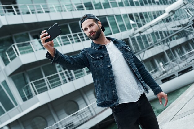 スマートフォンの笑顔でselfie写真を撮って街の通りに立っているアウトドアレジャー若いスタイリッシュな男