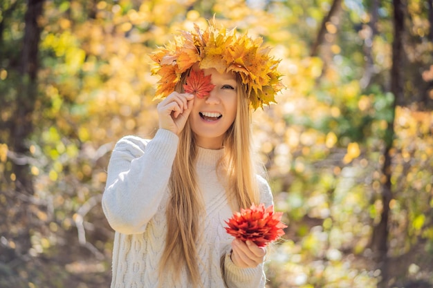 Outdoors lifestyle close up portrait of charming blonde young woman wearing a wreath of autumn leaves Smiling walking on the autumn park Wearing stylish knitted pullover Wreath of maple leaves