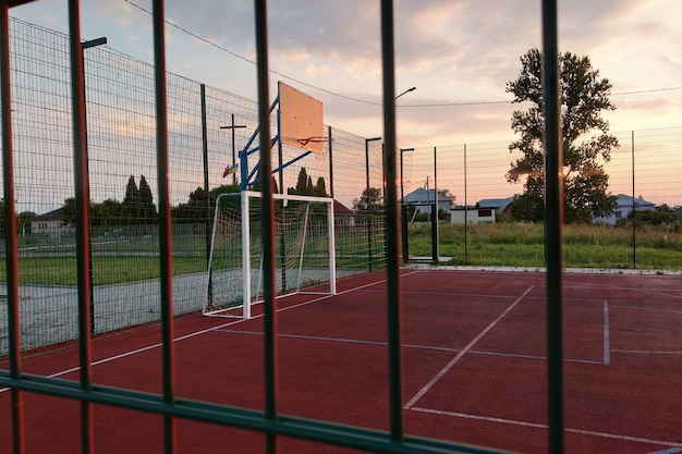 На открытом воздухе площадка для мини-футбола и баскетбола с воротами для мяча и корзиной, окруженной высоким защитным забором.