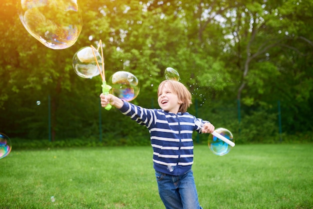 Outdoors portrait of cute preschool boy blowing soap bubbles on a green lawn