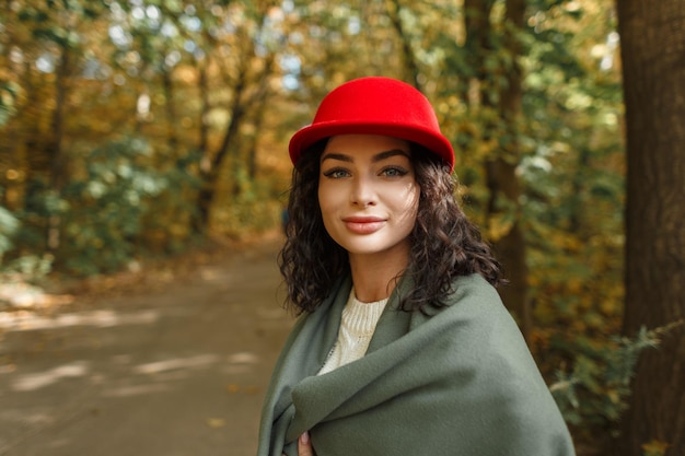 가을 공원에서 빨간 모자와 니트 스웨터가 달린 스카프가 달린 패션 옷을 입은 행복한 아름다운 세련된 소녀의 야외 초상화