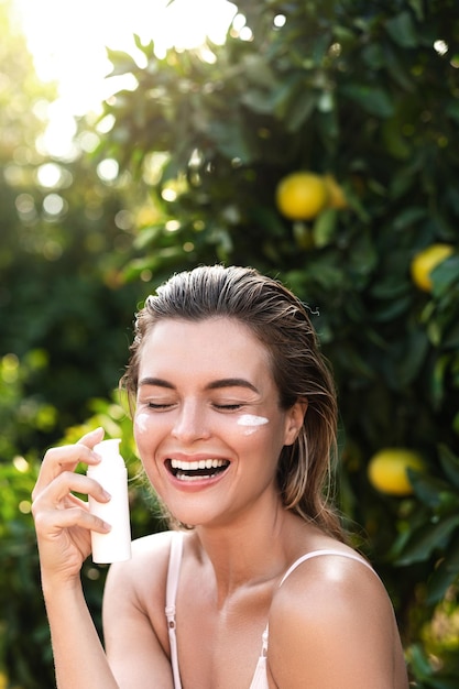 Outdoors portrait of joyful and beautiful woman applying moisturizing cream or sunblock on her facial skin