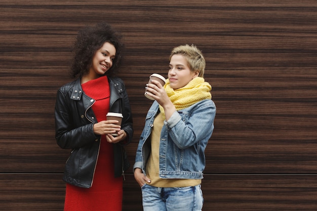 Photo outdoors portrait of two female friends. girls in casual warm outfits having city walk in cold season, drinking coffee and posing at wooden background, copy space. urban lifestyle, friendship concept