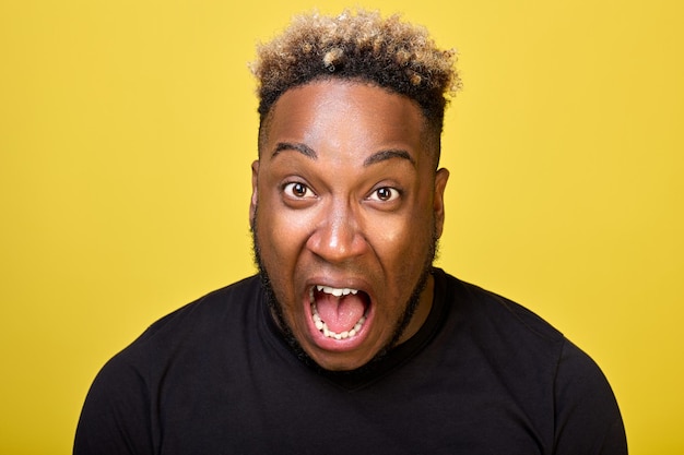 An outraged AfricanAmerican yells with all his might in outrage closeups on camera on a yellow background An enraged black guy screams loudly with his mouth wide open and bulging eyes