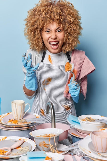 Outraged housewife screams loudly wears dirty t shirt apron\
protective rubber gloves holds brush washes up dishes stands near\
sink in kitchen busy doing housework during weekend dishwashing\
concept