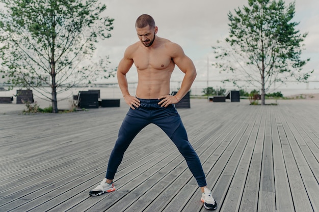 Outside of athlete guy does sport exercises in fresh air stands with naked torso has muscular body and stands outside Active unshaven guy in good physical shape has regular workout in morning