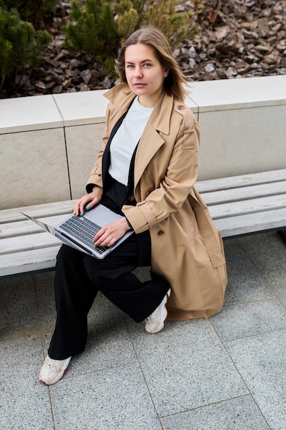 Outside the office businesswoman in a sleek trench coat takes a break enjoying the fresh air while attending to her laptop