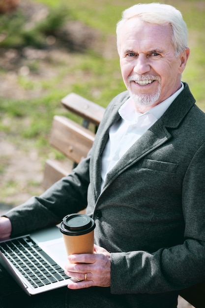 Outside office. Jovial mature businessman using laptop while looking at camera