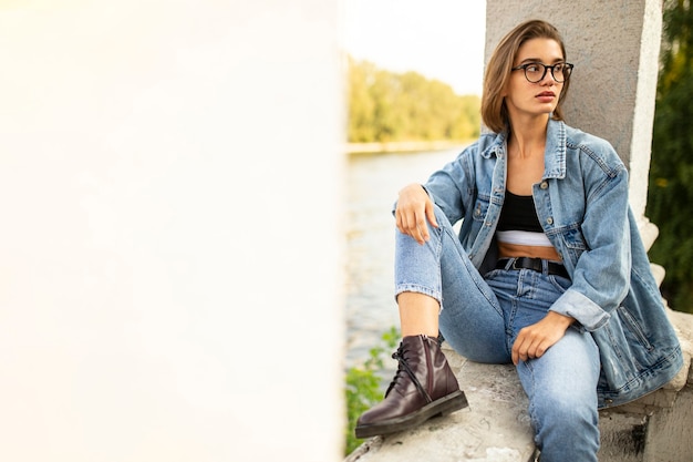 Foto fuori il ritratto di una giovane donna bionda hipster, vestita di jeans e stivali, seduta fuori