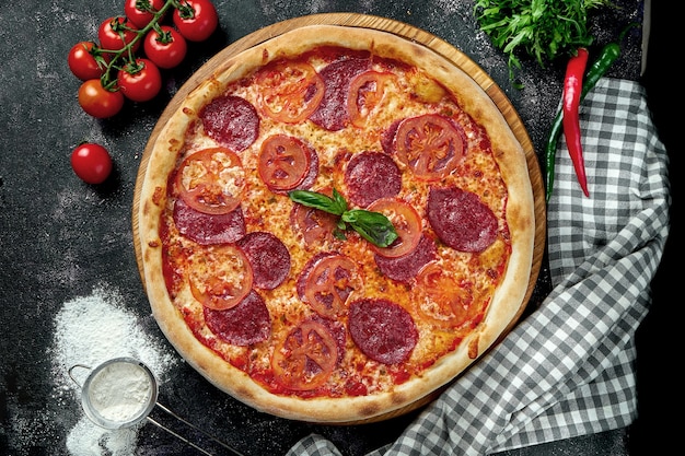 Oven-baked Italian pizza with red sauce, cheese, tomatoes and pepperoni salami in a composition with ingredients on a dark table. View from above.