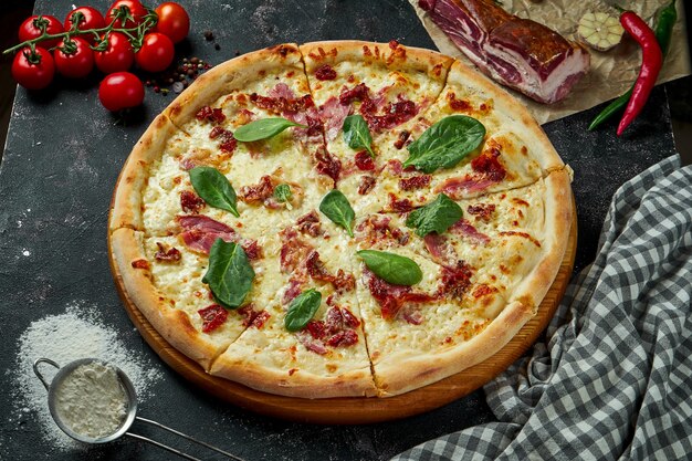 Запеченная в духовке итальянская пицца с соусом, сыром, ветчиной и вялеными помидорами в композиции с ингредиентами на темном столе. Закрыть вверх