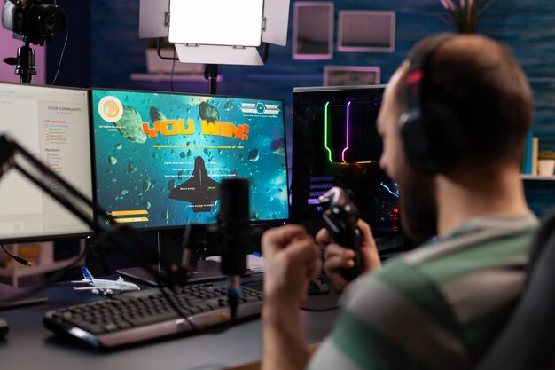 Over schouder beelden van professionele streamer die digitale videogames speelt op computer met koptelefoon, microfoon en bediening. Streaming Man steekt handen op voor het winnen van space shooter