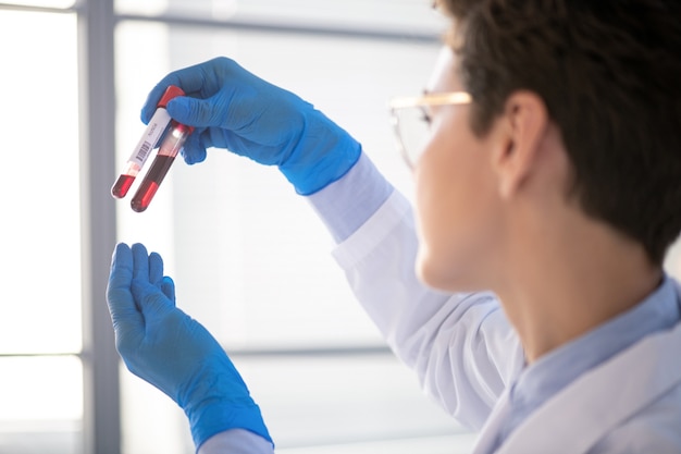 Over schouder weergave van vrouwelijke laboratoriumspecialist in handschoenen reageerbuizen op licht houden tijdens het onderzoeken van bloed door coronaviruspatiënt