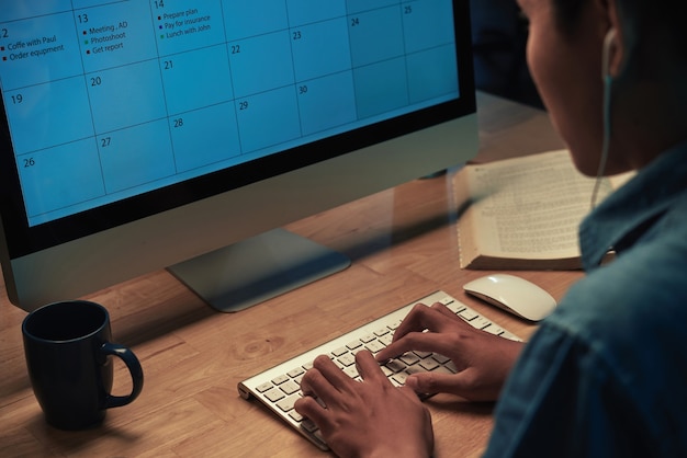 Over schouder zicht op aziatische man zit aan houten bureau met boek en mok en gebruikt online kalender o...