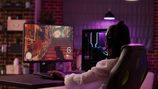 Over schouder zicht van Afro-Amerikaanse gamer meisje die online games speelt met behulp van gaming pc-setup die thuis ontspannende tijd doorbrengt. Vrouw die moderne technologie gebruikt voor entertainment en streamingtoernooien.