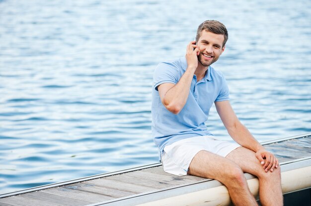 Overal verbonden blijven. Knappe jonge man in poloshirt praten op de mobiele telefoon en glimlachen terwijl hij op de kade zit