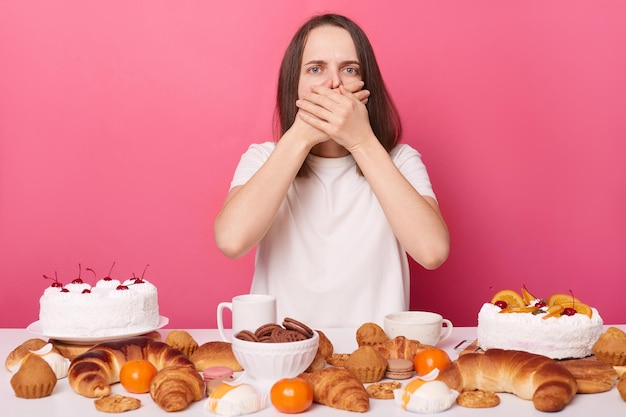 Overeating sad brown haired woman in white t shirt sitting at table isolated over pink background feeling nausea after eating sugary cakes