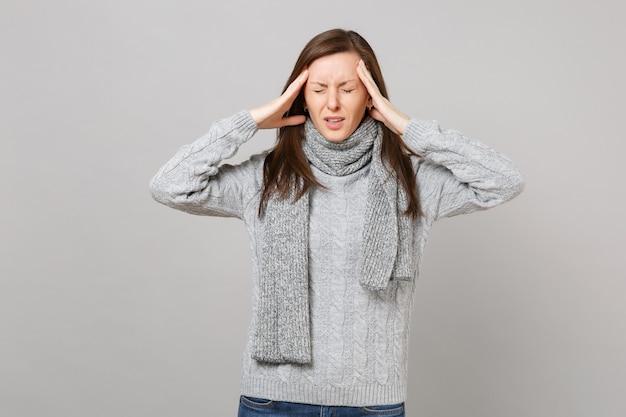 Overexhausted young woman in gray sweater, scarf with closed eyes putting hands on head, having headache isolated on grey background. Healthy lifestyle ill sick disease treatment, cold season concept.