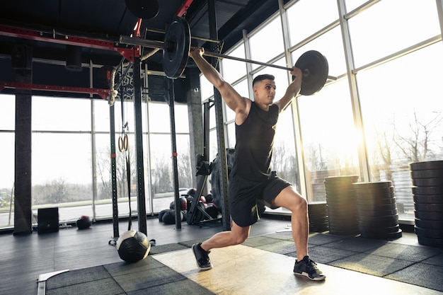 Overgaan. Jonge gespierde blanke atleet traint in de sportschool, doet krachtoefeningen, oefent, werkt aan zijn bovenlichaam met gewichten en barbell. Fitness, wellness, gezond levensstijlconcept.