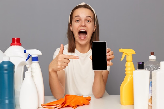 Overgelukkige blanke vrouw draagt hoofdband en casual t-shirt zittend aan tafel met verschillende wasmiddelen wijzend op smartphone met leeg scherm voor reclame geïsoleerd over grijze achtergrond