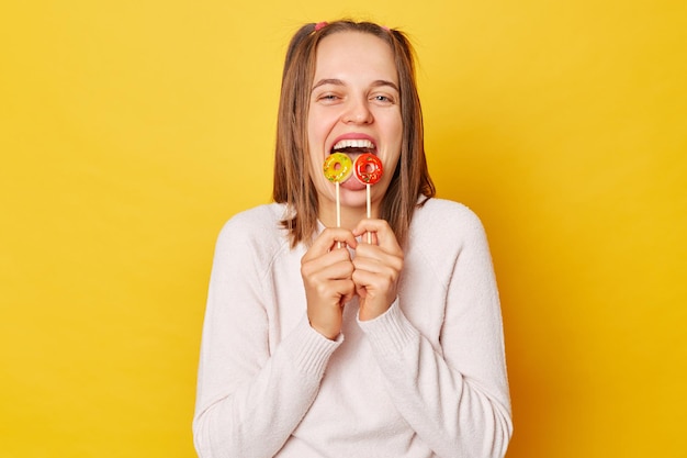 Foto overgelukkige suikerverslaafde vrouw met paardenstaart in trui met lolly geïsoleerd over gele achtergrond likken aan snoepjes verheugd om desserts te eten