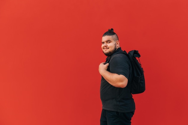 overgewicht jonge man in stijlvolle vrijetijdskleding staat met een rugzak op zijn rug tegen rode muur