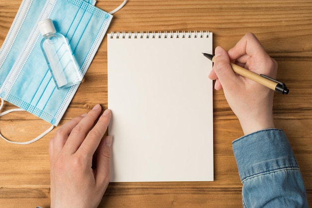 Overhead blank notebook hands with pen blue mask and bottle of antiseptic isolated on the wooden backdrop