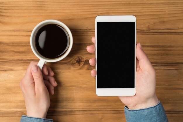 Overhead foto van smartphone in handen en kopje koffie geïsoleerd op de houten achtergrond