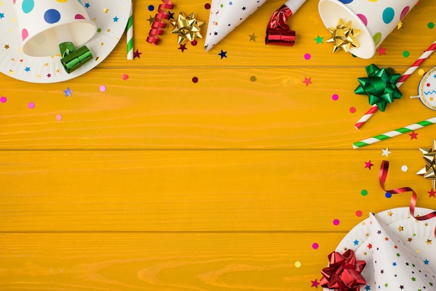 Overhead photo of cups drink with party tubules whistle multicolored confetti bow candle birthday hat and plates isolated on the yellow wooden background with empty space