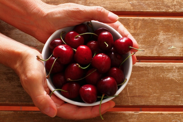 overhead photo of hands holding up a white bowl with cherries on wooden slatted table