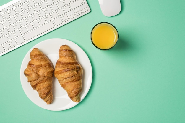 Foto aerea della tastiera del mouse del computer bicchiere di succo e piatto con due croissant isolati sullo sfondo verde acqua