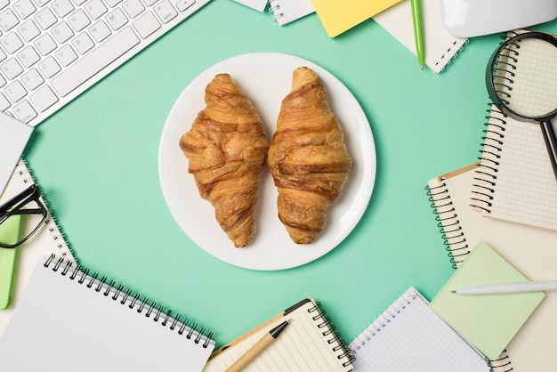 Photo overhead photo of keyboard computer mouse magnifier pens glasses two croissants in the middle with plate and heap of notebooks isolated on the teal backdrop