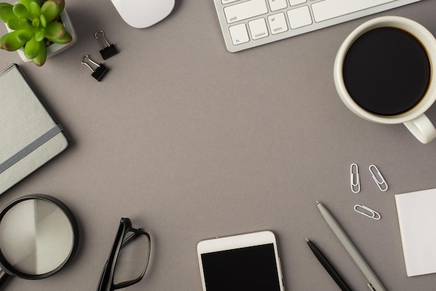 Overhead photo of notebook keyboard computer mouse magnifier pens phone plant glasses cup of coffee and paperclips isolated on the grey background with blank space