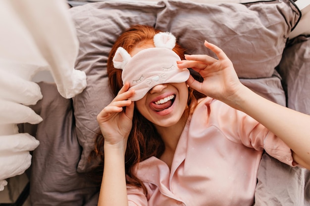 Overhead portret van lachende vrouw in roze nachtmasker Ginger Kaukasisch meisje chillen in bed