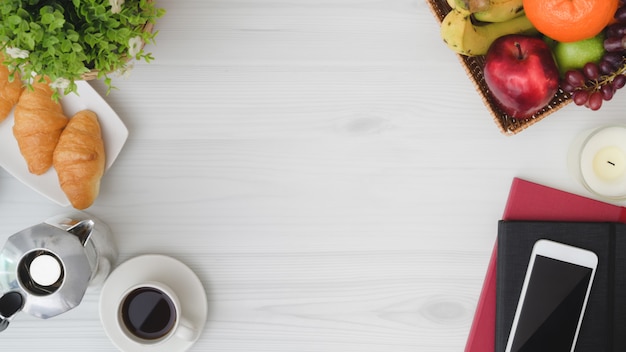 Overhead shot of  breakfast table with copy space, phone, notebook, fruit basket, croissant, coffee cup and moka pot