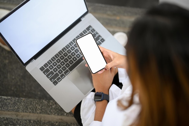 Overhead shot Female using smartphone and working on laptop computer white screen mockup