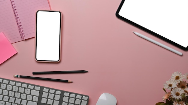 Overhead shot of female workspace with smart phone, digital tablet, notebook and keyboard on pastel pink background.