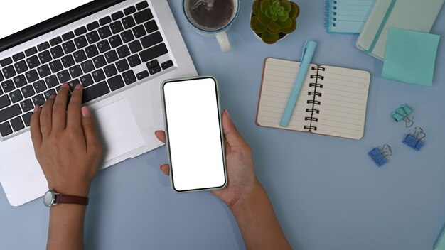 Overhead shot of woman holding smart phone and working on laptop computer on pastel blue surface