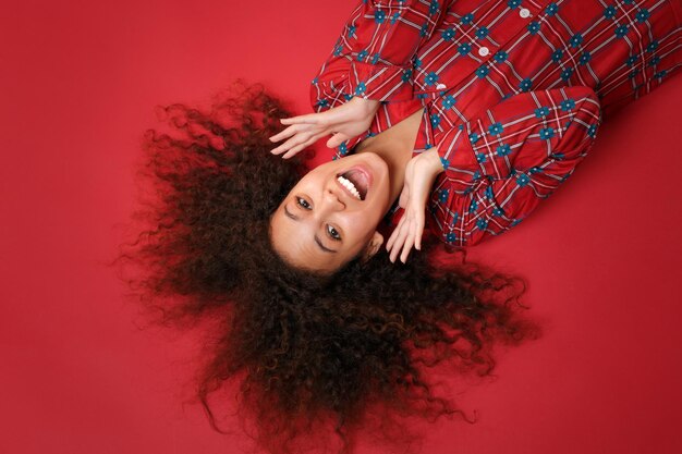 Overhead top view of excited young african american girl in pajamas homewear resting at home isolated on red background. Relax good mood lifestyle concept. Lying, keeping mouth open, spreading hands.