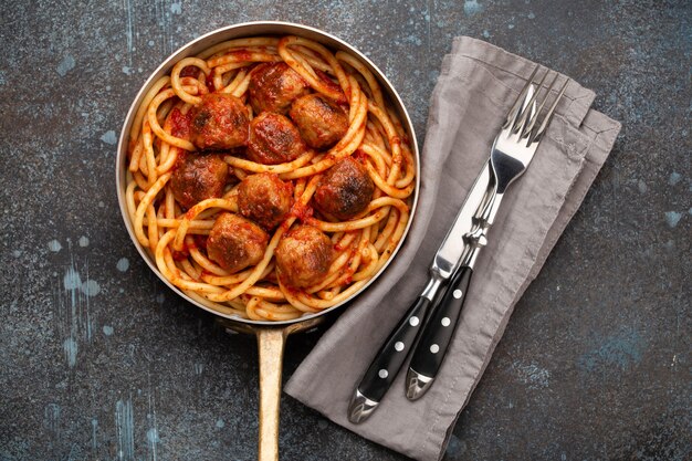 Overhead of traditional homemade pasta with roasted meatballs, tomato sauce in copper vintage pan with fork and knife on rustic concrete background. Classic homemade meatballs spaghetti for dinner