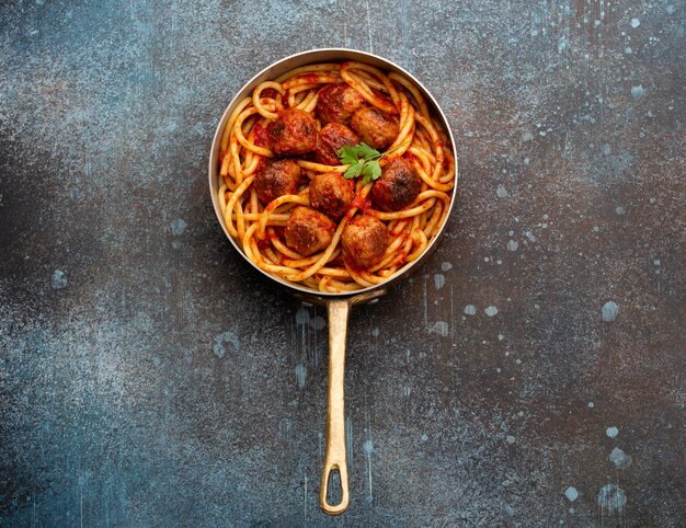 Overhead of traditional homemade pasta with roasted meatballs, tomato sauce, green herbs in copper vintage pan on rustic concrete background. Classic homemade meatballs spaghetti for dinner