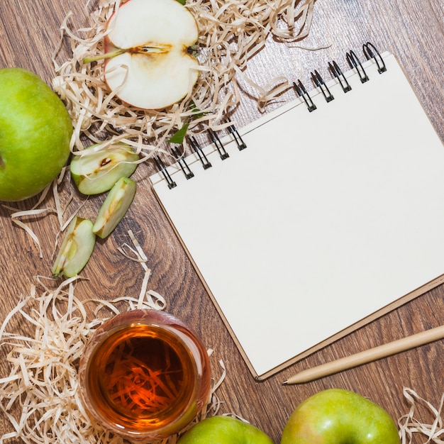 An overhead view of apple cider vinegar; green apples and spiral notepad with pencil on wooden desk