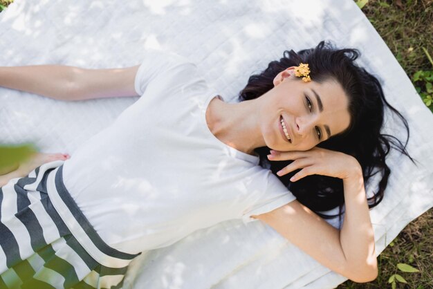 Overhead view of beautiful cheerful young Caucasian woman smiling and looking at the camera lying on white blanket on the grass with flower on hair Wellness relaxation travel and people concept