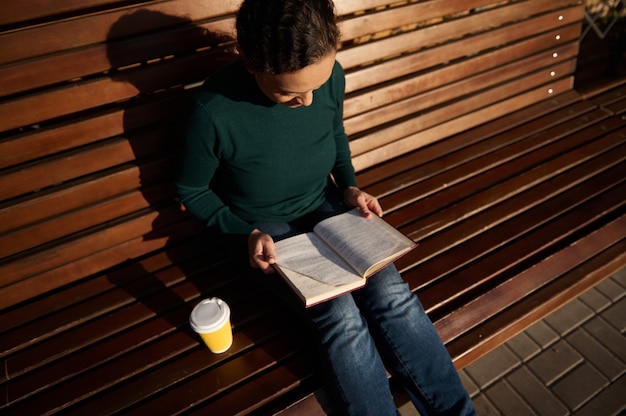 Overhead view of beautiful young brunette sitting on wooden bench in park, reading book and drinking coffee. Serene confident woman in casual denim with a book and takeaway paper cup of hot drink