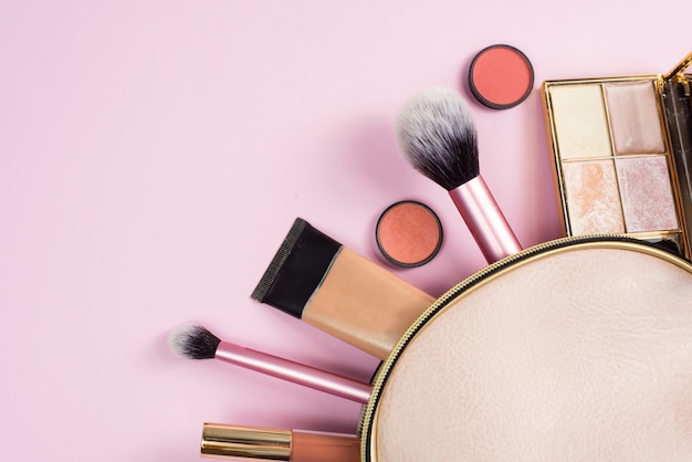 Overhead view of a beige leather make up bag, with cosmetic beauty products spilling out on to a pastel pink background