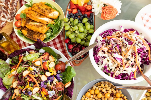 Overhead view of bowls of summer salads, bread and fruit, ready to eat on an outdoor table decorated with flowers. Family enjoying time at home, lifestyle concept