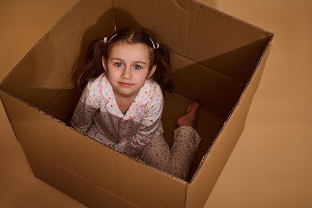 Overhead view of a charming little European girl with two ponytails wearing a pajamas with colorful dots sitting inside a cardboard box and looking at camera isolated over beige background
