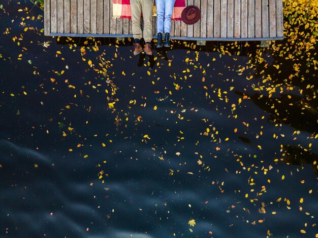 Overhead view of couple legs at the wooden pier lake water with yellow leaves