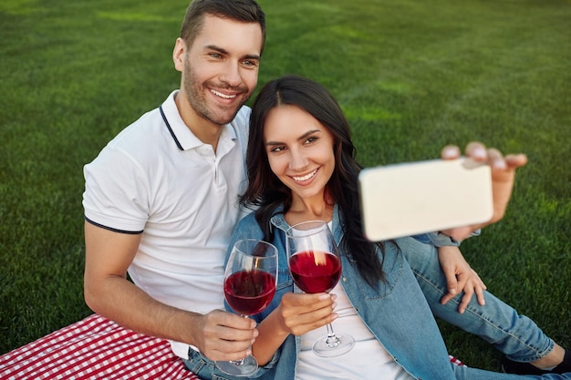 Вид сверху на пару, делающую селфи на смартфоне в парке, держащую бокалы с красным вином